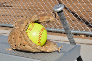 photo of softball mitt with a softball and bat on a dugout bench.