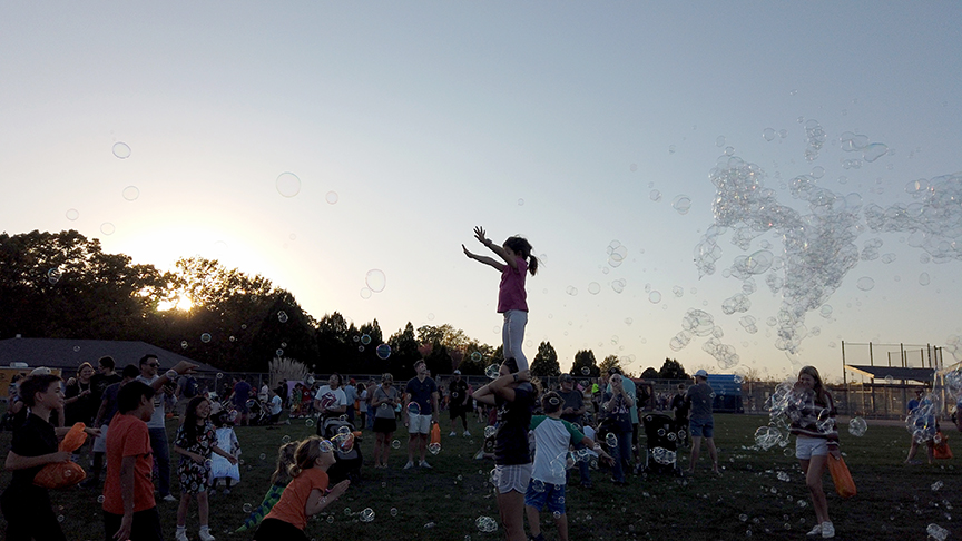 A crowd chases after huge bubbles with one girl standing on her mom's shoulders reaching for a bubble.