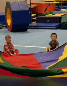 Two young boys smile while holding on to a huge parachute in the gymnastics center.