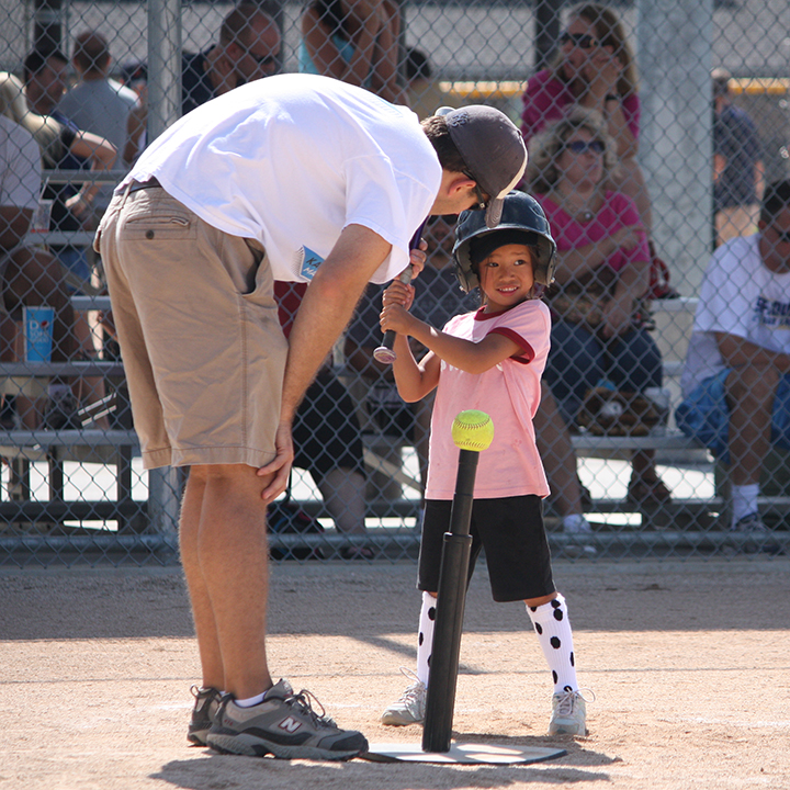 Coach helping a young girl hit a softball off a tee at home plate.