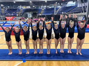 Young gymnasts raise their hands while standing in a line during a gymnastics meet.