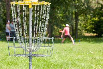 Disc golf basket with a player throwing in the background.