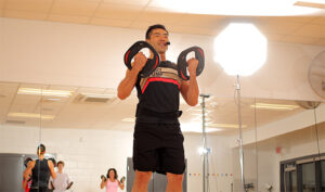 An instructor shouts out commands while holding Les Mills barbells.