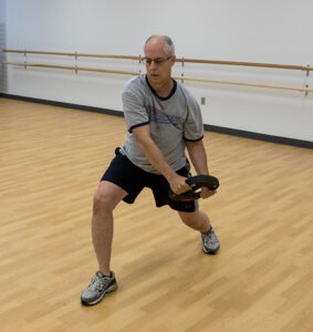 A man squats with legs apart while twisting a weight to his side.