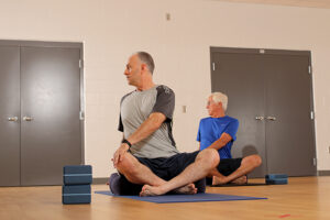 Two men sit in a cross-legged position while turning their torso to stretch.