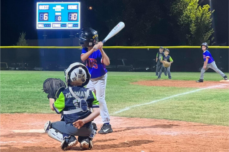 Youth baseball player at bat during a nighttime game while the catcher and pitcher prepare for the next pitch.