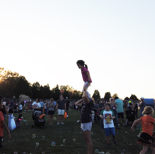 Children and families gathered outdoors at a community event while a child is lifted into the air above the crowd.